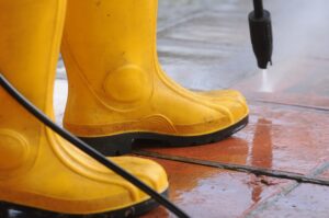 Person wearing yellow rubber boots with high-pressure water nozzle cleaning the dirt in the tiles